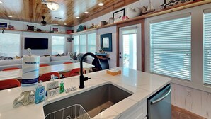 A modern kitchen with a marble countertop, a sink, and a dishwasher. There are various kitchen items on the counter, including a bottle of soap and a sponge. The kitchen has wooden cabinets and a window with blinds.