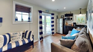 Una sala moderna con un sofá y un sillón, una cocina con refrigerador y microondas, y una ventana con persianas.