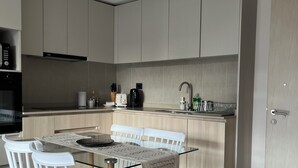 A modern kitchen with a dining area. It includes a glass-top table, white chairs, and wooden legs. There is a built-in oven and a coffee machine on the counter.