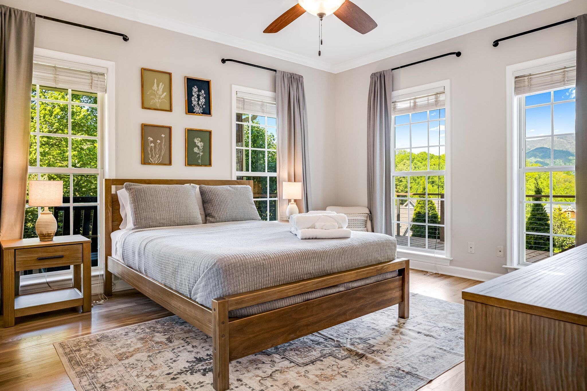 A bedroom with a wooden bed, a nightstand, a bedside table, and a view of greenery through large windows.