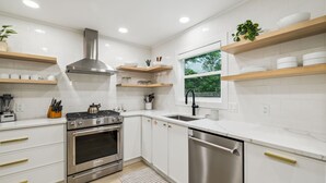 A modern kitchen with white cabinets, stainless steel appliances, wooden shelves, and a window providing a view of greenery outside.
