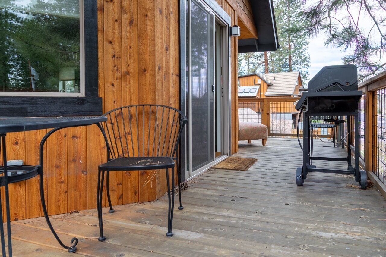 A wooden deck with a grill and chairs, a sliding glass door, and a view of a wooden building in the background.