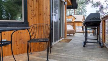 A wooden deck with a grill and chairs, a sliding glass door, and a view of a wooden building in the background.