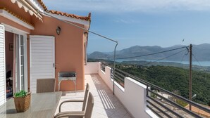 Un balcón con una silla y una mesa de mimbre, una puerta blanca y vistas a montañas y un lago.