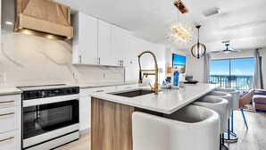Eine moderne Küche mit weißen Schränken, einem eingebauten Backofen, einer großen Kochinsel mit Barhocker und Meerblick durch große Fenster.