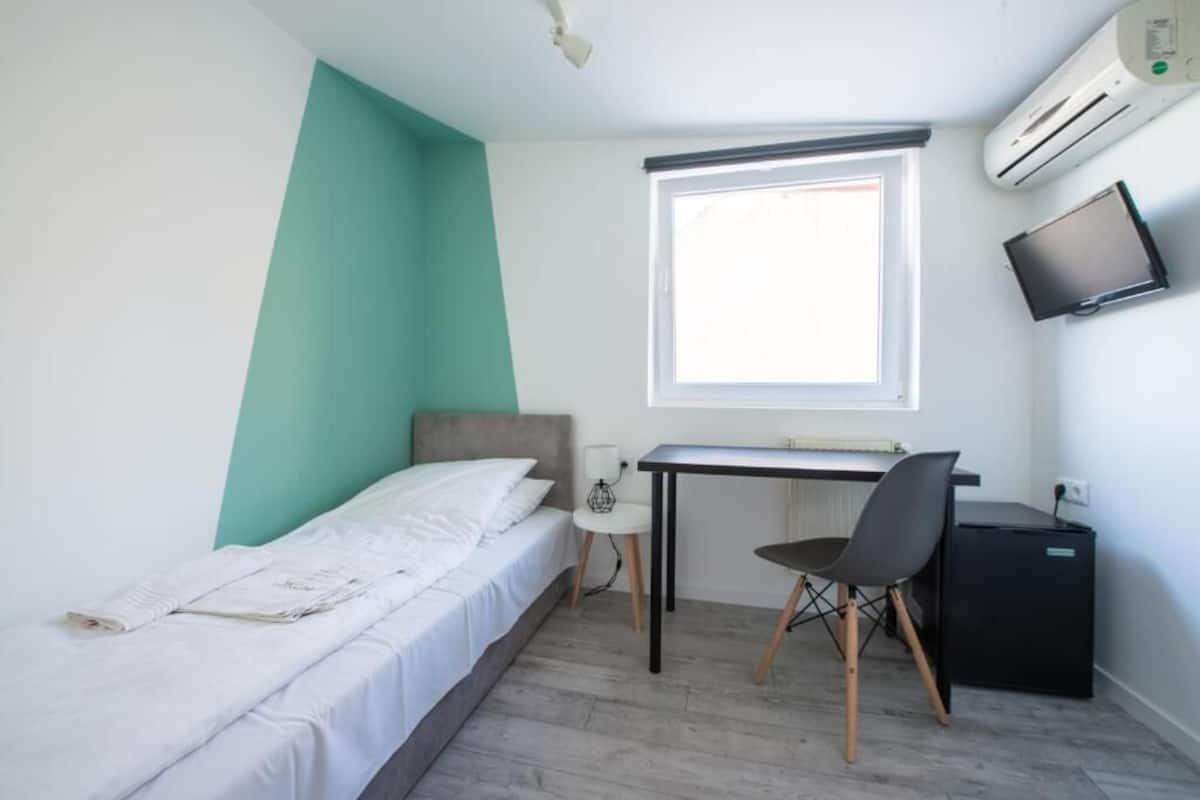 A compact room with a bed, a desk, a chair, a small table, a TV, and an air conditioner.