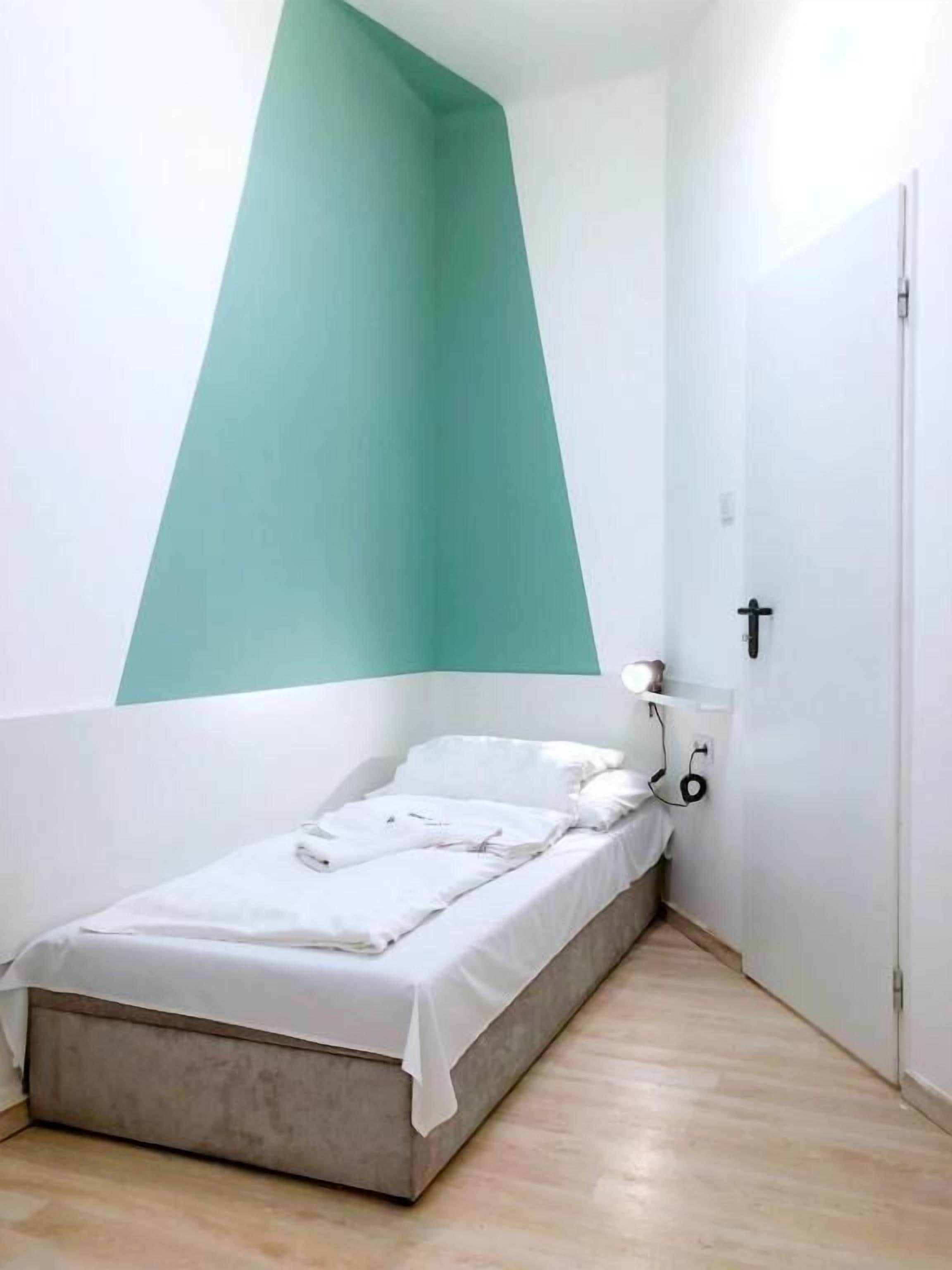 A small, neatly arranged bedroom with a bed, a bedside table, and a wall-mounted light fixture.