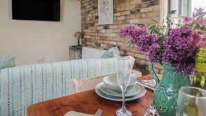 A dining area with a brick wall, a wooden table set for two, and a green vase with purple flowers. - Drake's Hideaway (Lyme Regis)