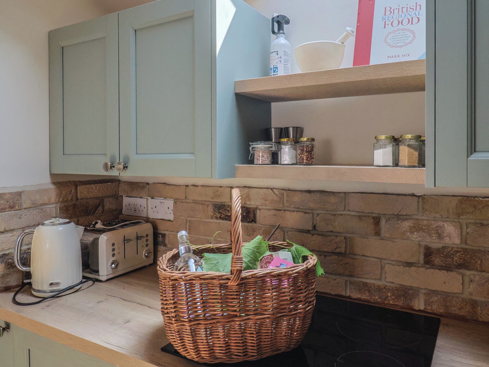 A kitchen with light green cabinets, a brick wall, a toaster, a kettle, a wicker basket with green leaves, and a shelf with jars and a book titled "British Regional Food".