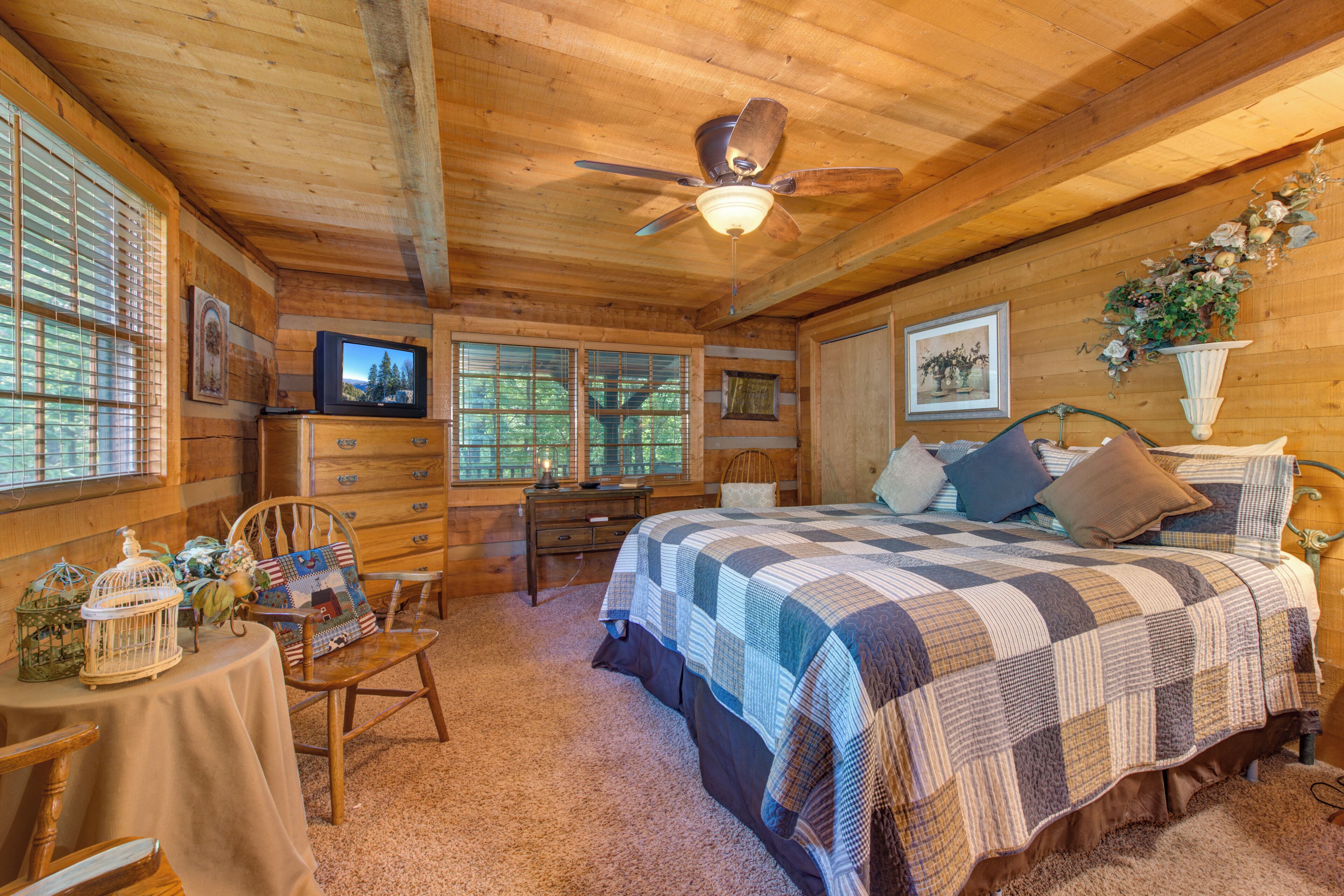 A cozy log cabin bedroom with a bed, a television, a wooden dresser, and a window with blinds.