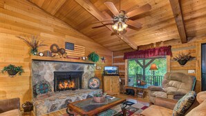 A cozy cabin interior with a stone fireplace, wooden walls, a ceiling fan, and a comfortable seating area.