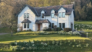 Une grande maison à deux étages, à l’extérieur blanc et au toit foncé, entourée d’un jardin bien entretenu où poussent différentes sortes de fleurs et où s’étend une pelouse.