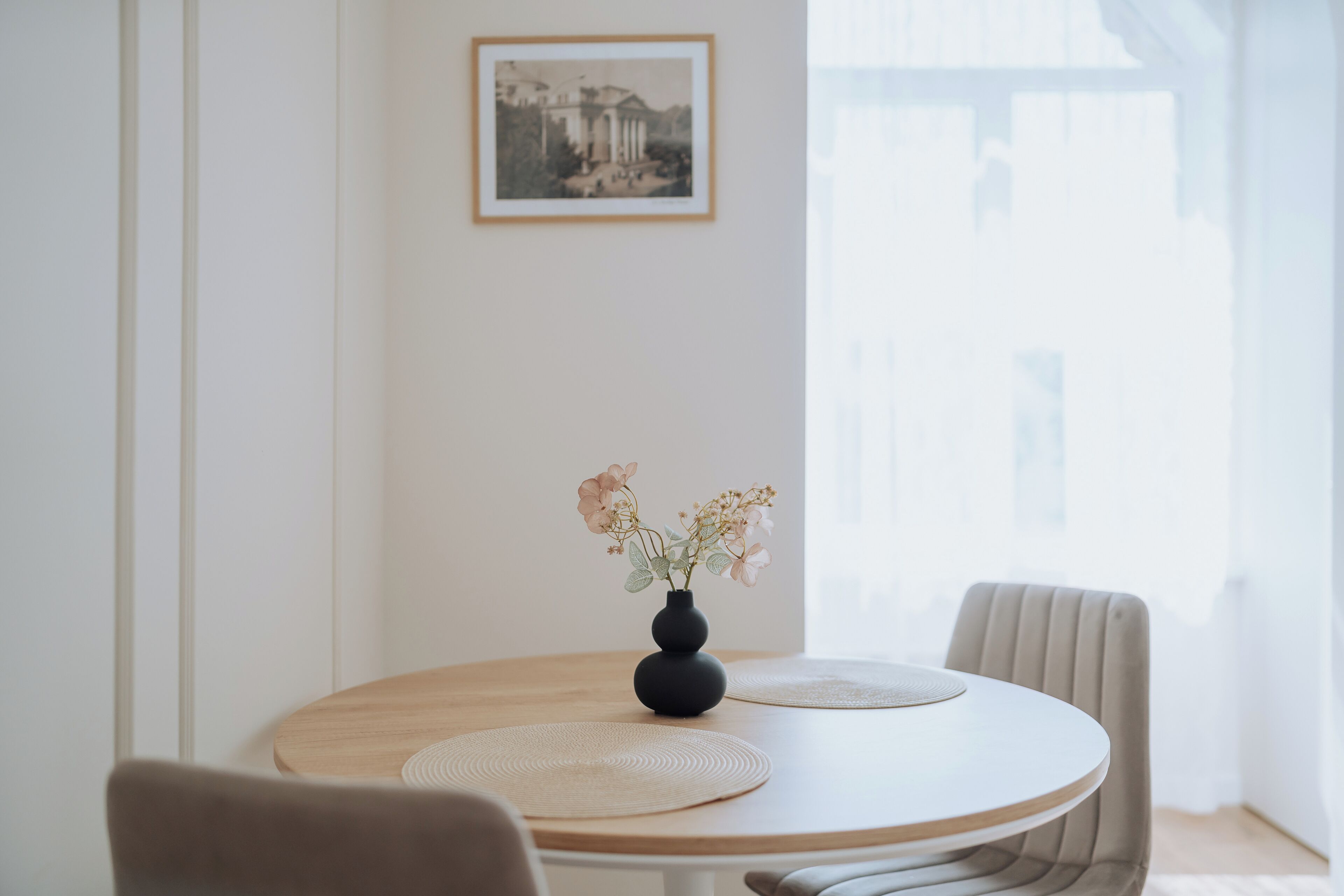 Una mesa redonda de madera con dos individuales beige y un florero oscuro con flores.