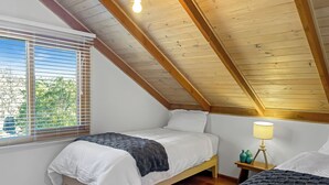 Een slaapkamer met een schuine kap, houten balken, een bed met wit beddengoed, een nachtkastje met een lamp en een raam met jaloezieën.