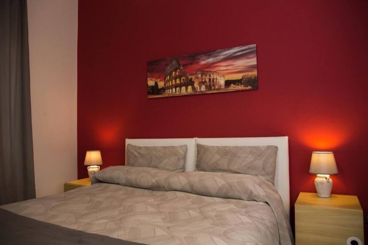 Une chambre d’hôtel avec un lit, deux lampes de chevet, un mur rouge et un tableau représentant un bâtiment historique.
