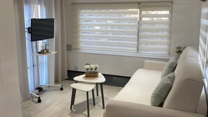 Ein modernes Wohnzimmer mit einer Couch, einem kleinen Tisch, einem an der Wand befestigten Fernseher und einem Fenster mit Jalousien.