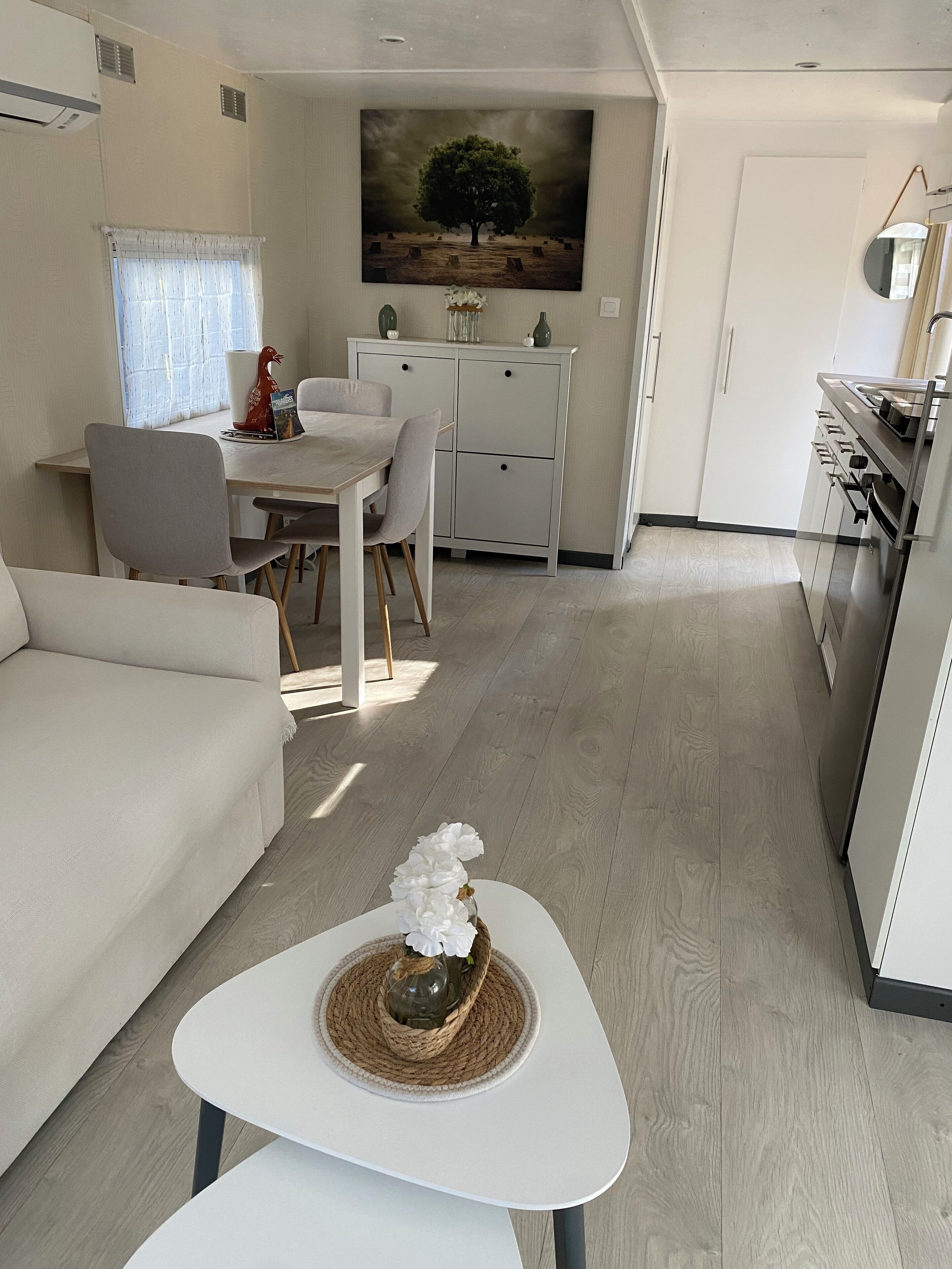 A modern living room with a sofa, a dining table with chairs, a kitchen area with cabinets and a stove, and a painting of a tree on the wall.
