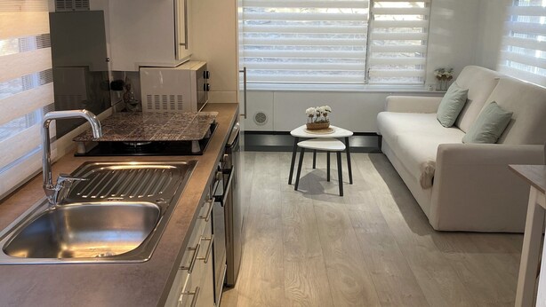 Eine moderne Küche mit Spüle und Herd aus Edelstahl, einer weißen Arbeitsplatte und Holzboden. Im Wohnbereich befindet sich eine weiße Couch und ein kleiner Tisch mit einer Blumenvase.