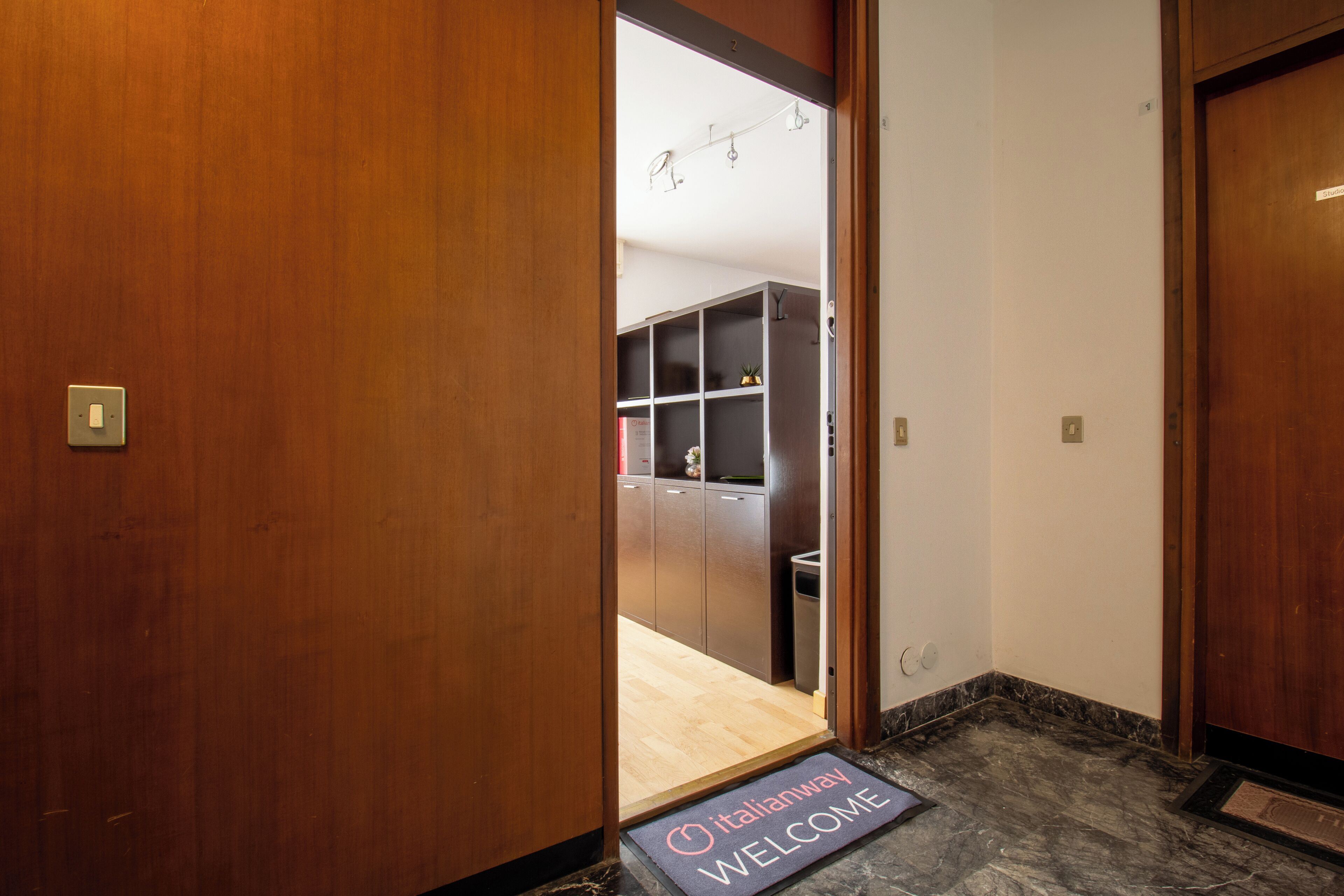 Een hotelentree met een houten deur, een welkomstmat en zicht op een kamer met kitchenette.