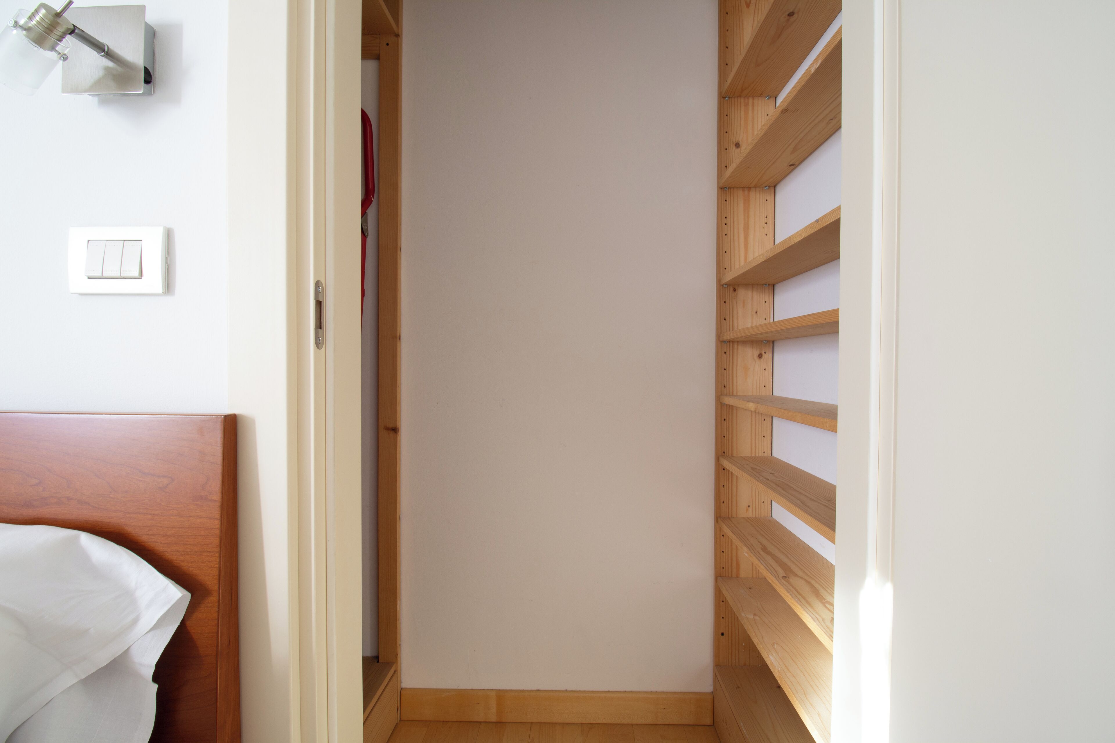 A narrow hallway with wooden shelving on one side and a bed with a white headboard on the other.
