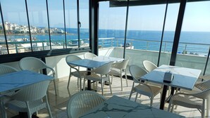 A rooftop dining area with white wicker chairs and marble tables, overlooking a coastal cityscape. - Aykaç Otel Susanoğlu (Silifke)