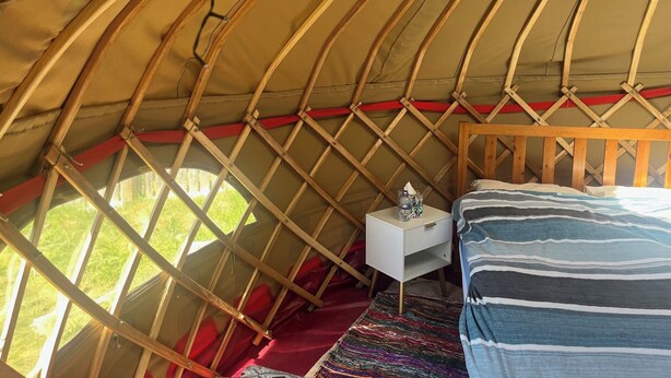 Een gezellig interieur van een joert met een houten bed, een klein nachtkastje en een kleurrijk vloerkleed.