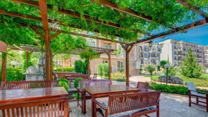 An outdoor patio with wooden tables and benches, surrounded by greenery and a view of apartment buildings. - Marino Mar | Boutique Apartment | Spa & Wellness, pools, sun loungers included (Sunny Beach)