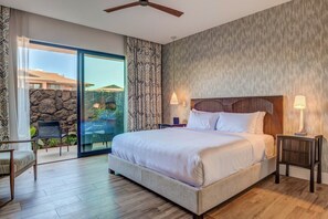 Un dormitorio con una cama grande, un ventilador de techo, una mesita de noche con lámpara, una silla y vistas a un patio con pared de piedra y plantas.