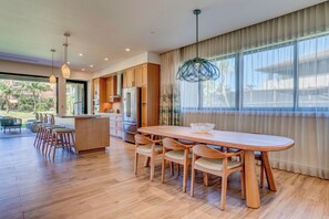 Una cocina moderna con armarios de madera, un comedor con una mesa grande y sillas, y ventanas amplias con cortinas ligeras.