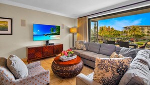 A living room with a sofa, armchairs, a coffee table, a TV, and a balcony with a view of buildings and greenery. - Honua Kai | Ocean View 6 BR, Sleeps 16 | Car Incl. w/ 6+ Nights | HKH ML-1097 by KBM (Lahaina)