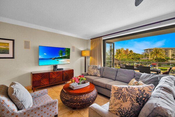 A living room with a sofa, armchairs, a coffee table, a TV, and a balcony with a view of buildings and greenery.
