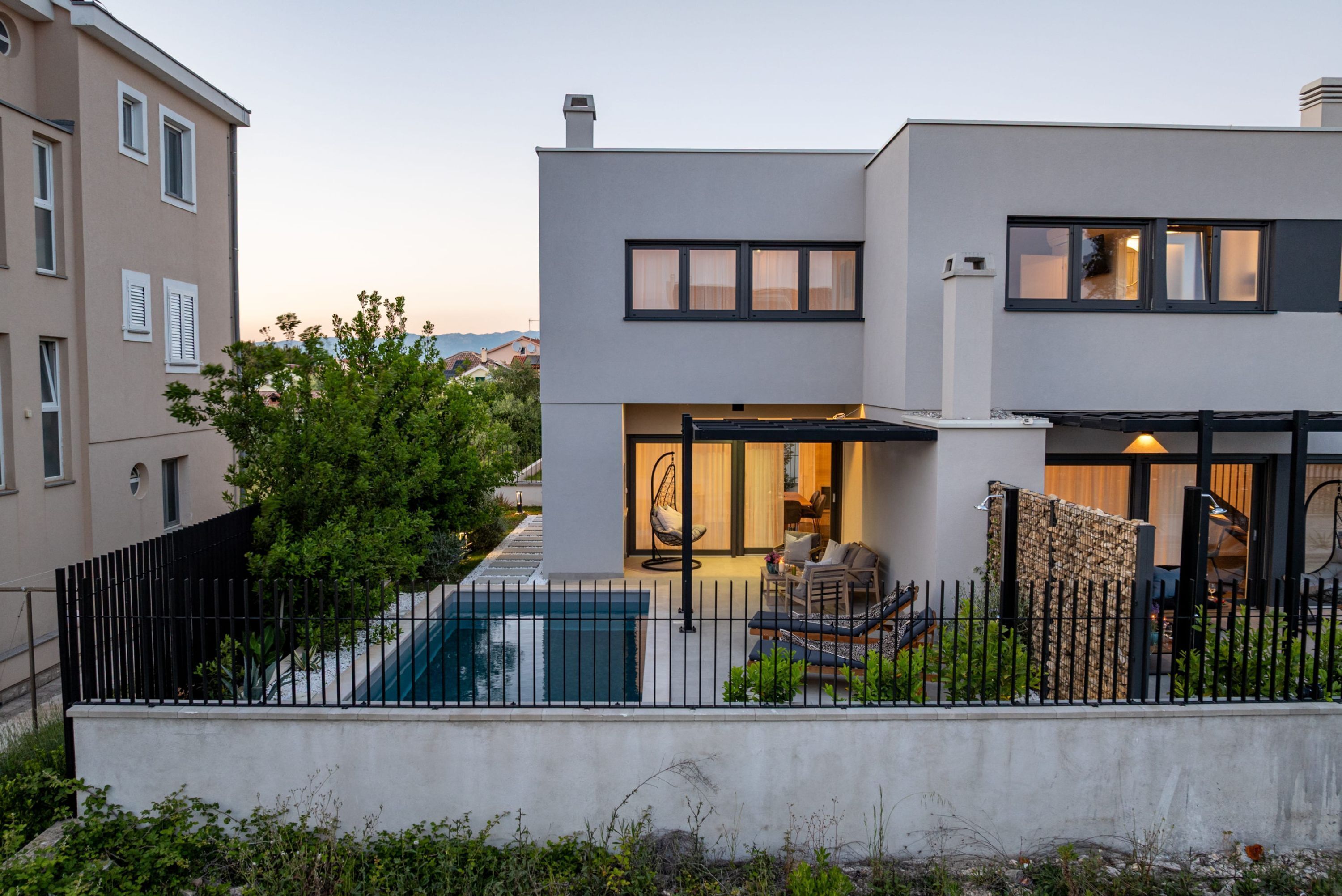 A modern two-story house with a swimming pool, a patio with outdoor furniture, and a garden area.