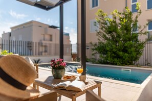 Escena junto a la piscina con una mesa, un libro, un vaso de jugo de naranja y un plato de comida.