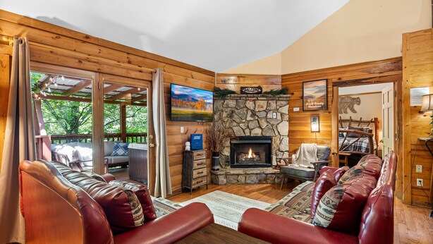A cozy cabin interior with a stone fireplace, leather sofas, a television, and a wooden deck visible through large windows.
