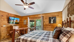 A log cabin bedroom with a plaid bedspread, a ceiling fan, a mounted TV, and a wooden chair.