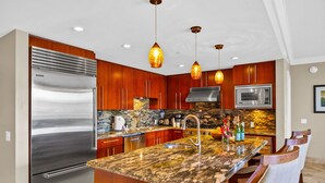 A kitchen with a stainless steel refrigerator, wooden cabinets, granite countertops, a microwave, and pendant lighting.