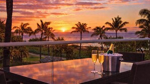 A table with two glasses and a bottle of champagne, overlooking a sunset with palm trees and the ocean.