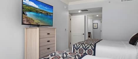 Quarto de hotel com televisão na parede, armário de madeira, duas camas com colchas estampadas e um ventilador de teto.