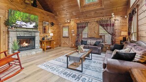 A rustic cabin interior with a stone fireplace, wooden walls, a large window, and a cozy seating area. - A Home For The Whole Family, Even The Family Dog (GATLINBURG)