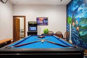 Een biljart met een groene bal, biljartkeus en een videogameapparaat in een ruimte met een muurschildering en een deur.