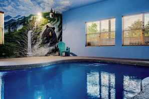 Une piscine ornée d’une fresque représentant un ours et des montagnes, une chaise verte et des fenêtres équipées de stores.