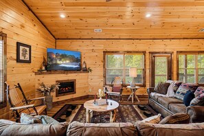 Un intérieur de chalet chaleureux avec une cheminée, des sièges confortables, un téléviseur et de grandes fenêtres.