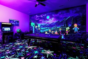 Une pièce avec une table de billard, des motifs d’étoiles au néon sur le sol, une fresque représentant des personnages animés et une borne d’arcade.