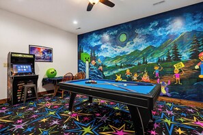 Une table de billard dans une pièce ornée d’une fresque murale, avec une console de jeux vidéo et un tapis à motifs étoilés.