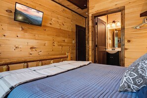 A wooden cabin bedroom with a bed, a television mounted on the wall, and a bathroom with a mirror and lights.