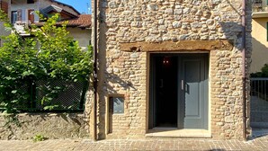 A stone building with a small window and a door, surrounded by greenery and other buildings. - La casa di Valbelluna (Feltre)