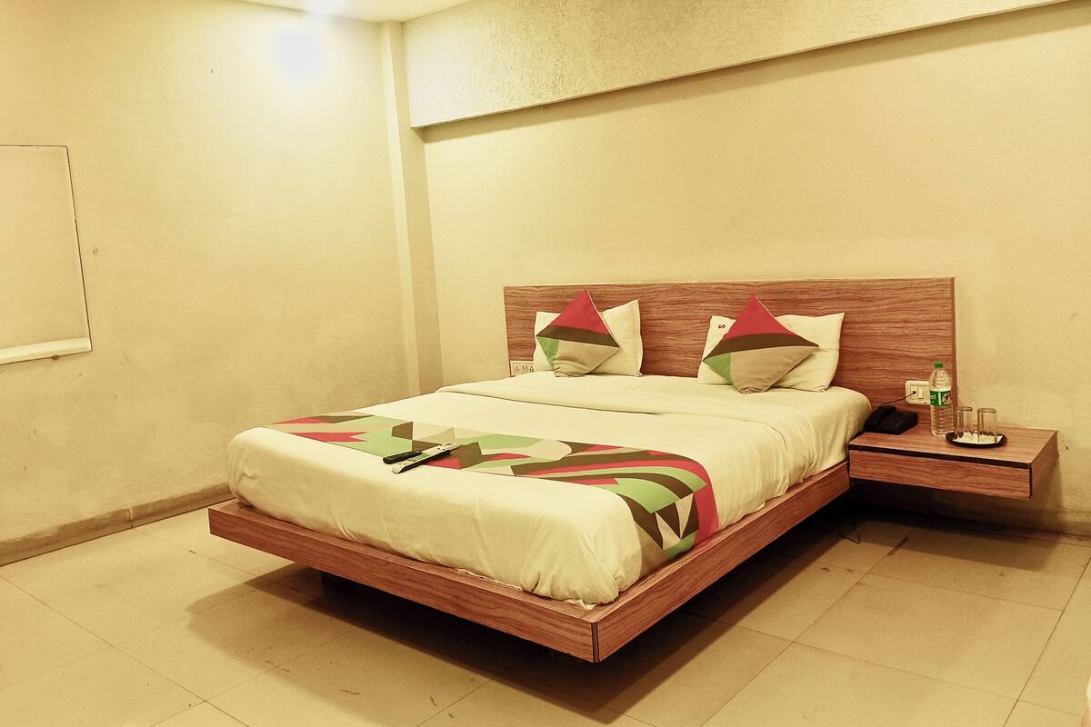 A modern hotel room with a wooden bed, a bedside table with a phone and glasses, and a wall-mounted light fixture.