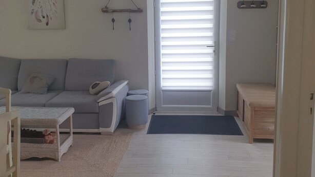 A living room with a grey sofa, a small table, and a bench. There is a door with blinds, a wall-mounted shelf, and a dreamcatcher.