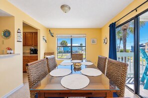 Un coin repas avec une table en verre et des chaises en osier, une cuisine avec des meubles en bois et une vue sur l’océan par de grandes fenêtres.