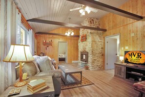 Una sala de estar acogedora con chimenea de piedra, paredes de madera, ventilador de techo y un letrero de MVR Vacation Rentals.
